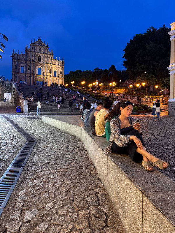 Люди сидят на каменных ступенях у исторического памятника в сумерках — городской релакс и наблюдение за прохожими в туристической зоне Макао.