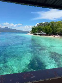 Кадр с лодки: прозрачная бирюзовая вода, мелководье у берега, песочный пляж, зелёные деревья и ясное голубое небо.