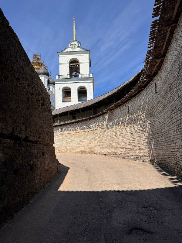 Каменная крепостная стена и белая башня внутри исторической части Пскова, узкая дорожка вдоль стены и чистое небо.