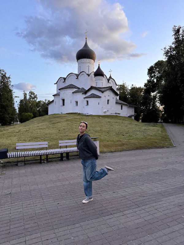 Белая церковь с куполом на зелёном холме и деревянной скамьёй на переднем плане; исторический архитектурный памятник Пскова.