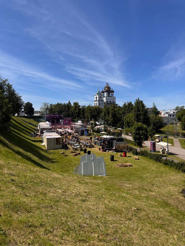 Парк у собора с зелёными склонами и временными торговыми палатками; летняя городская сцена и подготовка к локальному событию.