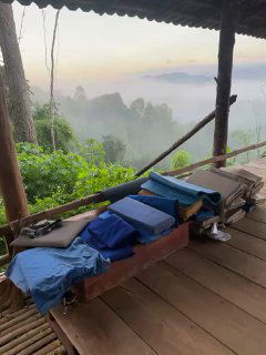 Палатка или место для отдыха с простыми лежаками на веранде, видно горный пейзаж и утренний туман в окружении леса.