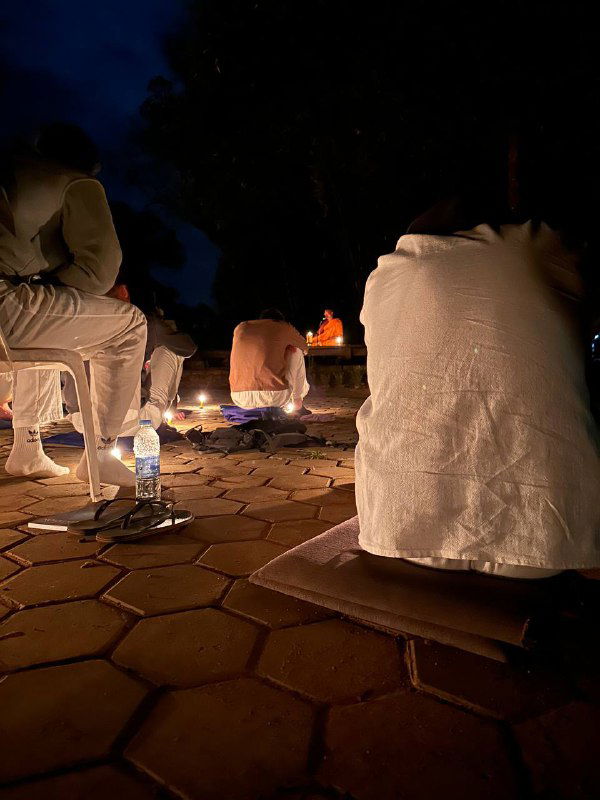 Ночная медитация на открытой площадке: люди в белой одежде сидят на матах при свечах и небольших фонариках, спокойная темная сцена.