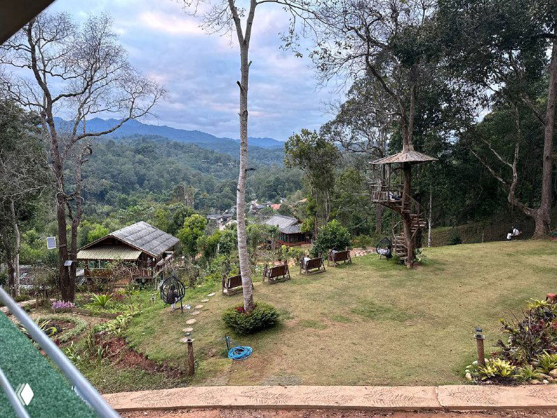 Панорамный вид с высоты на ретрит: озеленённый склон, несколько домиков и лесной массив на фоне гор и открытого неба.