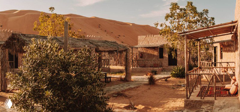 Панорамный вид на лагерь и деревянные постройки у подножия песчаных дюн; тёплая пустынная палитра и рассветный свет.