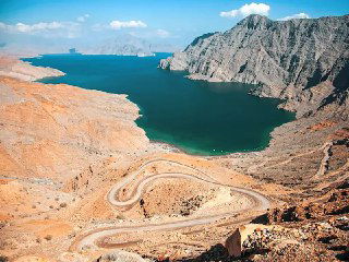 Извилистая дорога вдоль скалистых утёсов и узкий залив; виды на море и суровый прибрежный ландшафт с высоты.