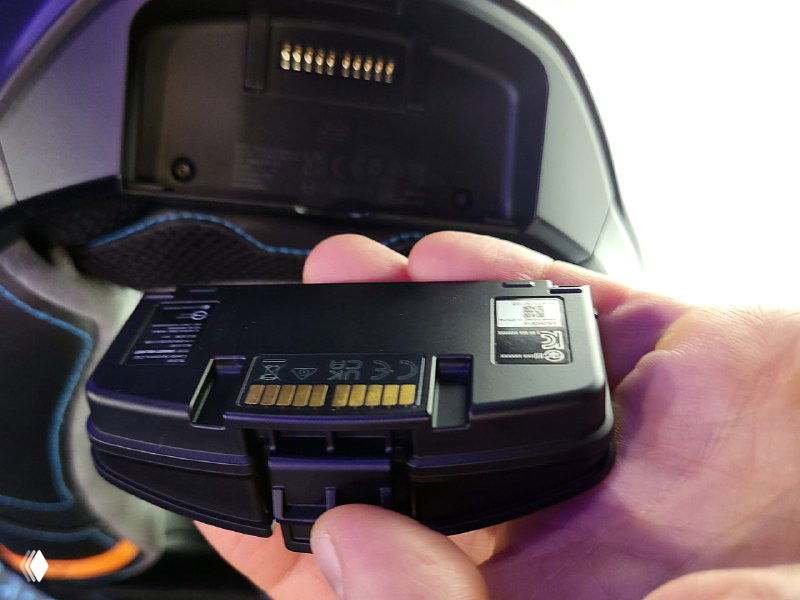 Рука держит отстёгиваемый модуль батареи и электроники шлема Cardo Beyond; видна контактная площадка, разъём и маркировка элемента питания.