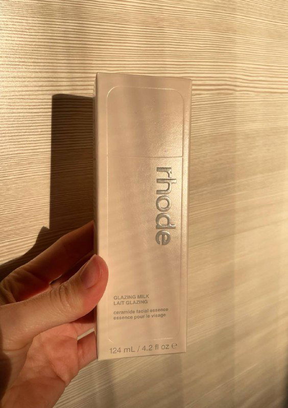 Упакованная картонная коробка Rhode в прозрачной плёнке и фирменным логотипом, рука держит упаковку на фоне светлой текстурной стены и тёплых бликов.