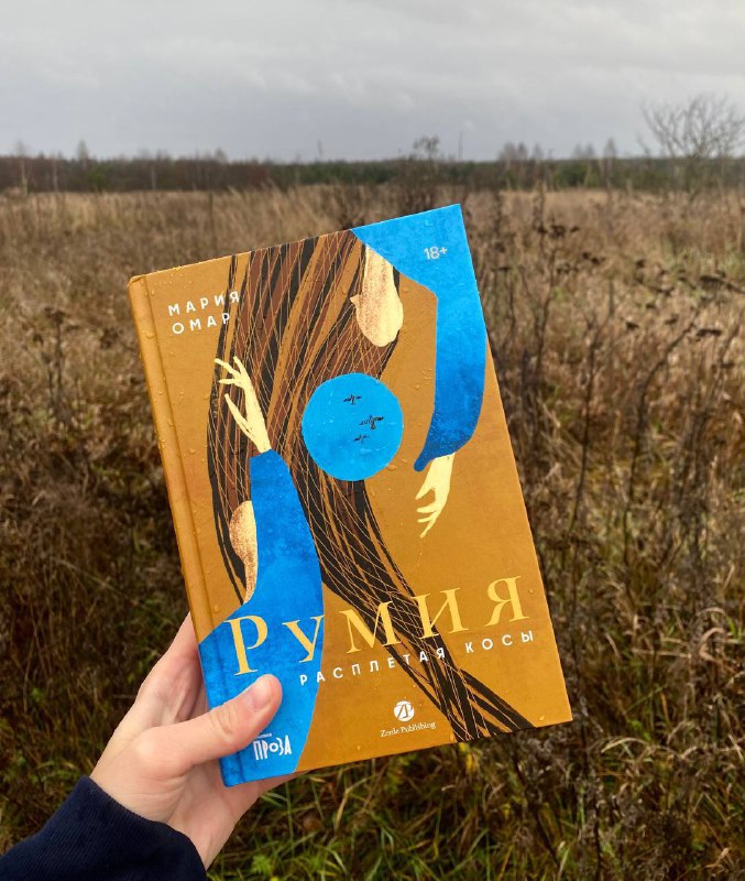 Рука держит книгу «Румия» Марии Омар на фоне осеннего поля; видна золотисто‑синяя обложка с заголовком и автором, создающая контекст чтения.