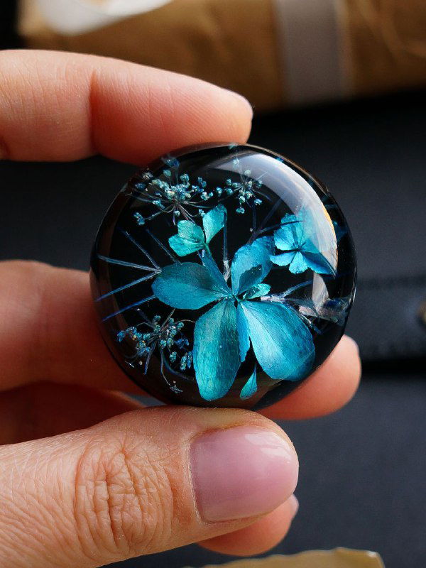 Круглая смолянная брошь с засушенными цветами голубых оттенков и тонкими стеблями зонтичного растения, держат пальцами.