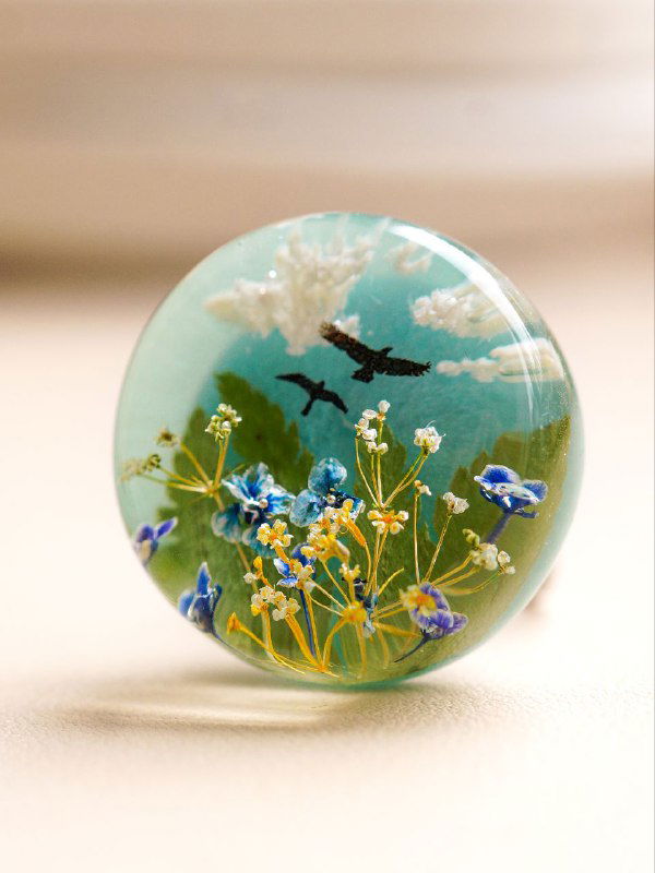 Круглый декоративный кабошон с миниатюрным пейзажем и микровставками засушенных зонтичных цветов на светлом фоне.