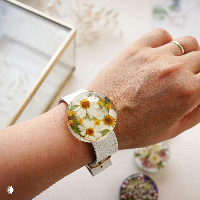 Наручные часы с широким белым ремешком и круглым смоляным циферблатом, внутри засушенные ромашки и зелёные элементы, на руке.