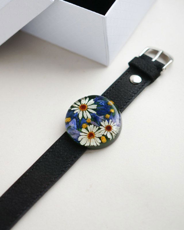 Чёрный кожаный ремешок с круглым смоляным украшением, внутри несколько белых ромашек на тёмно-синем фоне; минималистичная композиция.