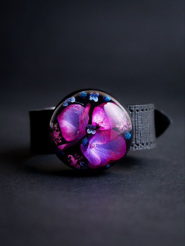Маленькая круглая брошь-кабошон с розово-фиолетовыми лепестками гортензии в прозрачной смоле, показана на матовом чёрном фоне.