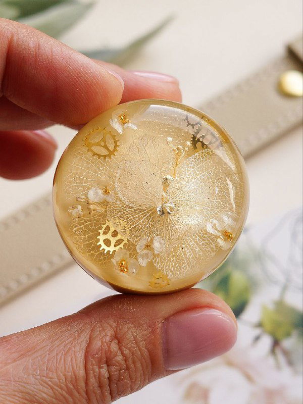 Круглый элемент с белыми и золотистыми деталями: фрагмент гортензии в прозрачной смоле, показан крупным планом в руке мастера для масштаба.