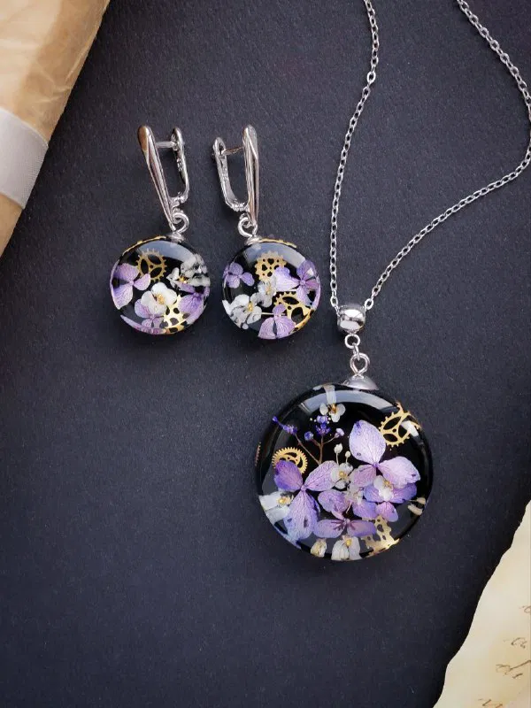 Комплект из серёжек и кулона с бледно-лиловыми высушенными гортензиями в прозрачной смоле, аккуратно выложенный на тёмном фоне для демонстрации комплектности.