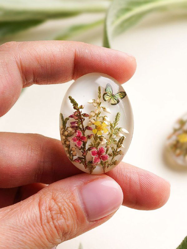 Овальный кабошон с небольшими полевыми цветами и алиссумом в прозрачной смоле, изображение демонстрирует масштаб изделия в руке