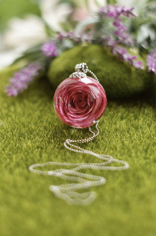 Колье с подвеской в виде круглой розы, композиция размещена на зелёной текстурированной поверхности, подчёркивающей цветок внутри смолы.
