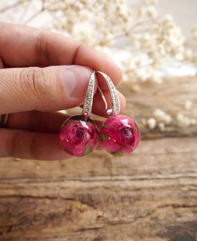 Пара подвесных серьг с круглыми смоляными шариками, внутри которых зафиксированы крошечные розовые бутоны; изделие на деревянной поверхности.