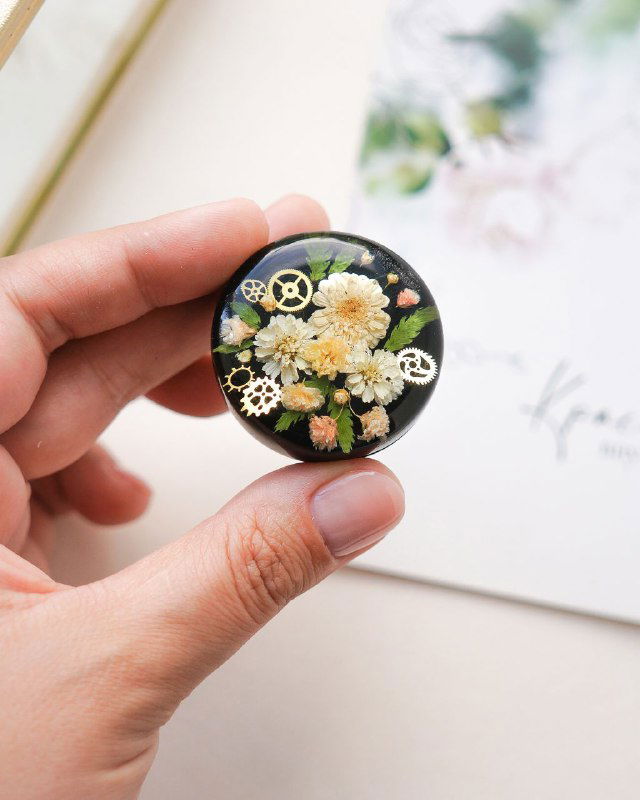 Круглая флористическая брошь/камейка на тёмном фоне с белыми цветами жемчужницы и декоративными металлическими шестерёнками в смоле.