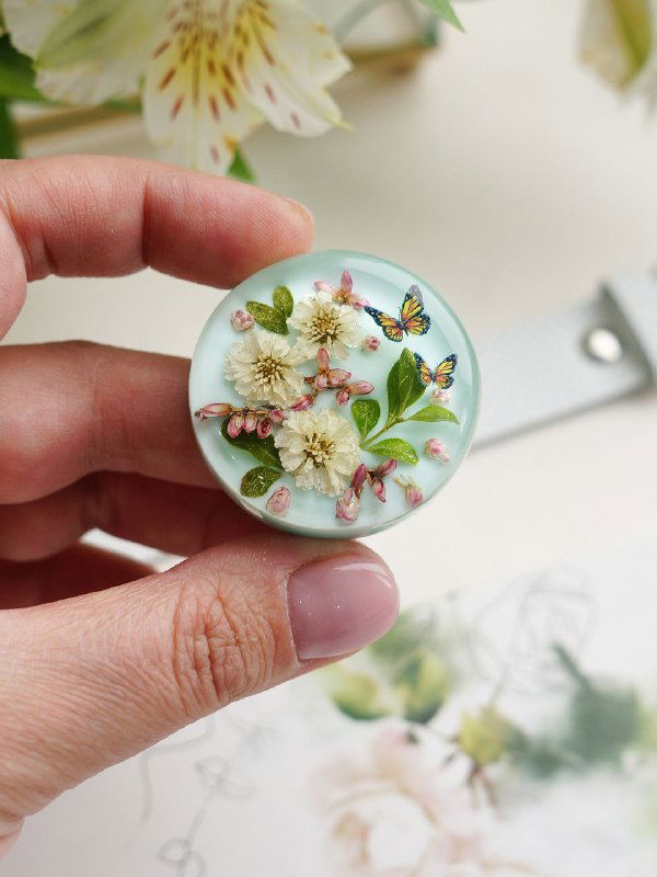 Крупный план круглого медальона на светлом фоне с мини‑цветами жемчужницы и декоративными элементами, видны детали и объём смолы.