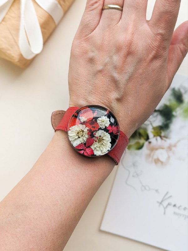 Рука в красном ремешке с круглым цветочным украшением из смолы, где живые бело‑красные композиции жемчужницы образуют яркий акцент.
