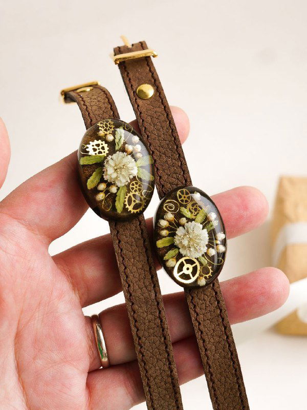 Две наручные ремешки коричневого цвета с круглыми флористическими вставками из смолы и белыми цветами жемчужницы, снимок в руке для масштаба.