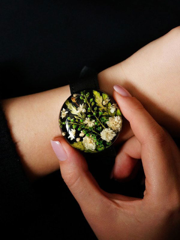 Темный фон и женская рука держат круглую подвеску с плотной зелёной композицией из множества мелких листьев и белых цветков.