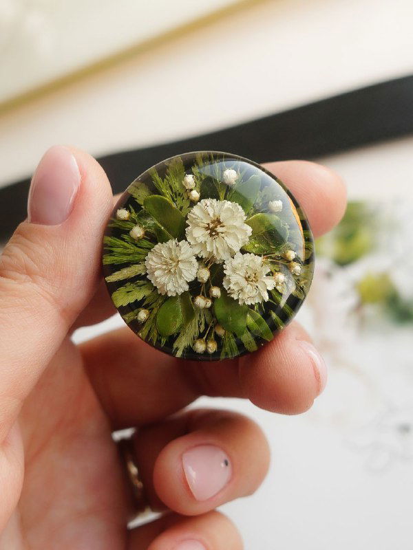 Круглая брошь в руке с букетиком белых соцветий и папоротниковых листьев, чёткая фактура просушенной зелени в эпоксидной заливке.
