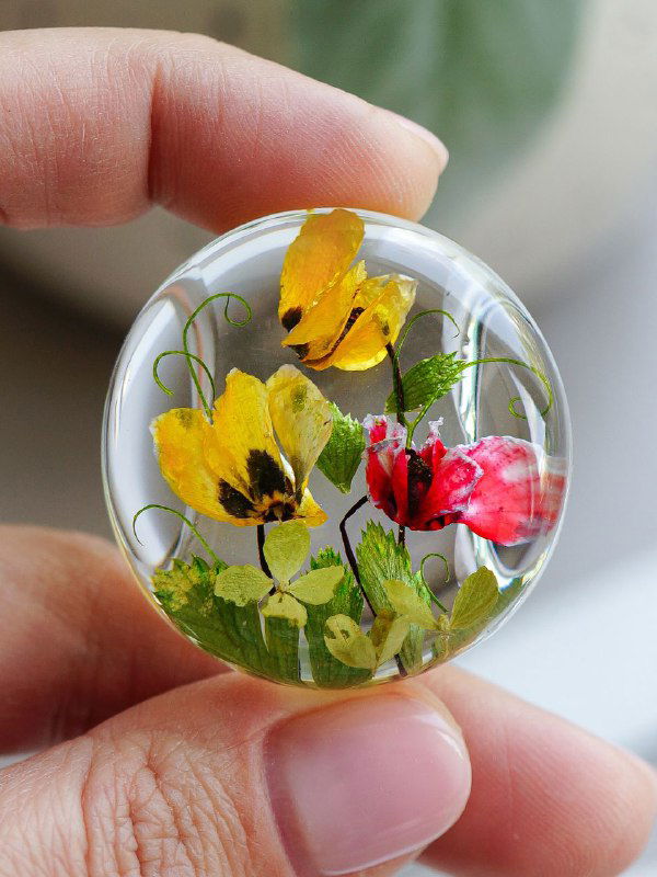 Крупный план прозрачного стеклянного кулона с яркими жёлтыми и красными цветами и зелёными стеблями, композиция выглядит натуральной и свежей.