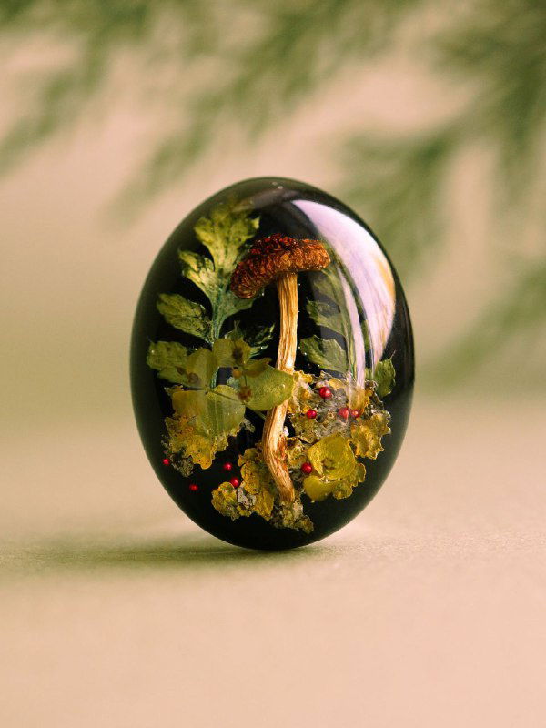 Овальная кабошон-подвеска из смолы с миниатюрной сушёной грибной композицией и зелёной листвой внутри на нейтральном фоне.