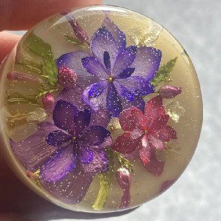 Круглый кабошон с пурпурными и розовыми цветами в прозрачной смоле; показана глубина, блеск поверхности и внутренняя текстура материала.