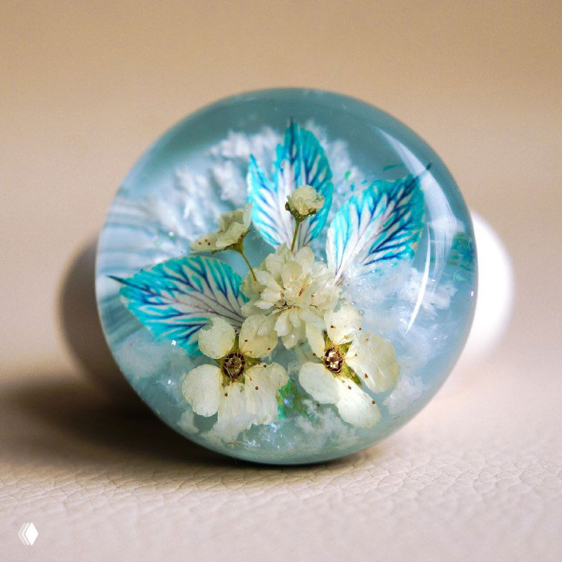 Круглый смоляной кабошон с белыми сухоцветами и бирюзово-голубыми акцентными листьями внутри на светлом фоне, детальный макро-кадр.