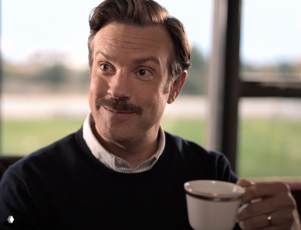 Портрет героя сериала Тед Лассо в тёплом свитере, улыбающийся и держащий чашку — образ тренера, внушающего доверие и оптимизм.