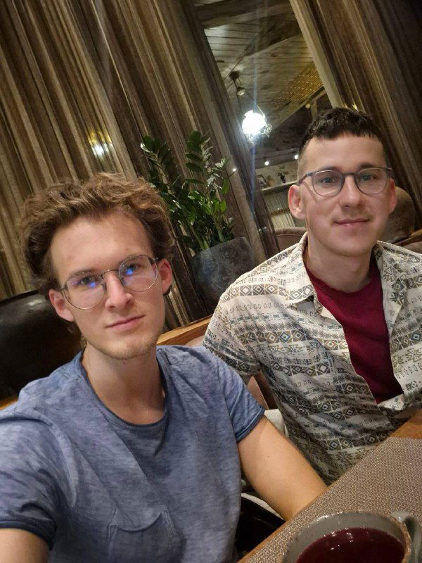 Два человека на фото в ресторане у окна, занавеси и растения на фоне, чашки на столе, спокойная обстановка и мягкий свет.