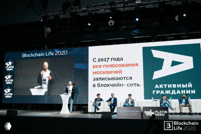 Широкий кадр сцены с большим экраном, где показан блокчейн-кейс и текст про голосование москвичей в блокчейн‑сеть; спикеры сидят и ведут дискуссию.