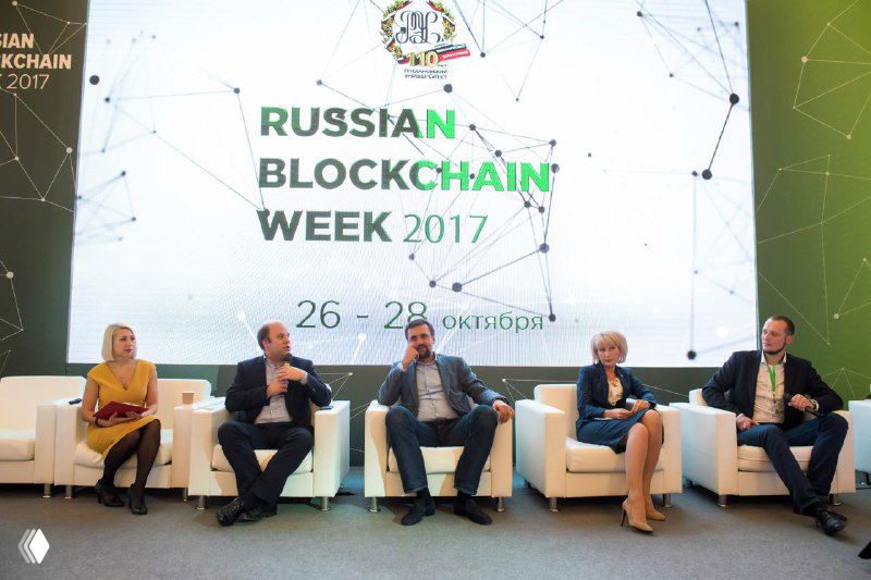 Панельные спикеры на сцене перед баннером Russian Blockchain Week — несколько человек сидят на белых креслах, конференц‑зона и брендирование на фоне.