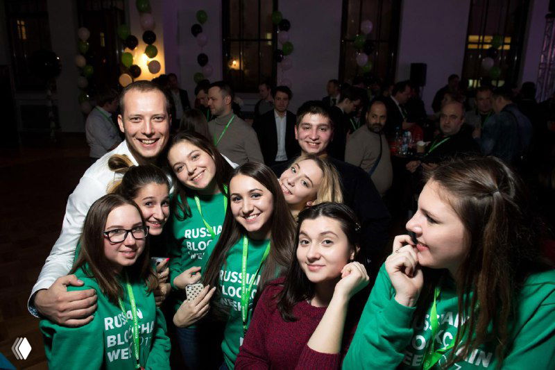Группа участников в зелёных футболках Russian Blockchain Week улыбается в камеру на фуршете — неформальное общение и атмосфера мероприятия после сессий.