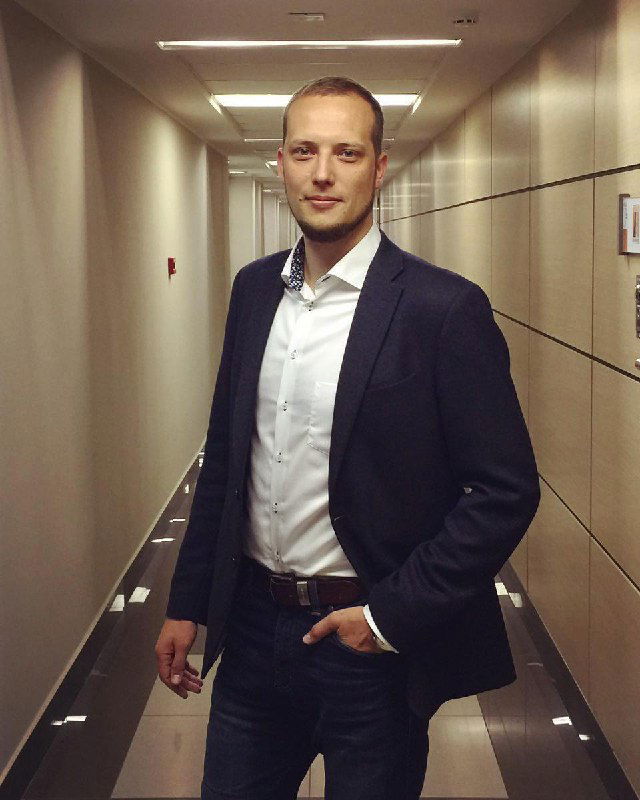 Портрет автора в коридоре офиса: мужчина в пиджаке и белой рубашке стоит в деловой позе, профессиональный биографический снимок для профиля