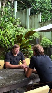 На фото двое мужчин сидят за большим каменным столом на открытой веранде; вокруг — пышная зелёная растительность и серая бетонная стена на заднем плане.