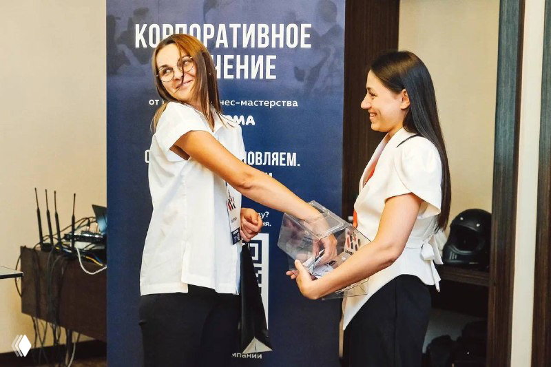 Девушка тянет руку в лототрон при розыгрыше подарков, вокруг видно внимание участников и организационные баннеры мероприятия.