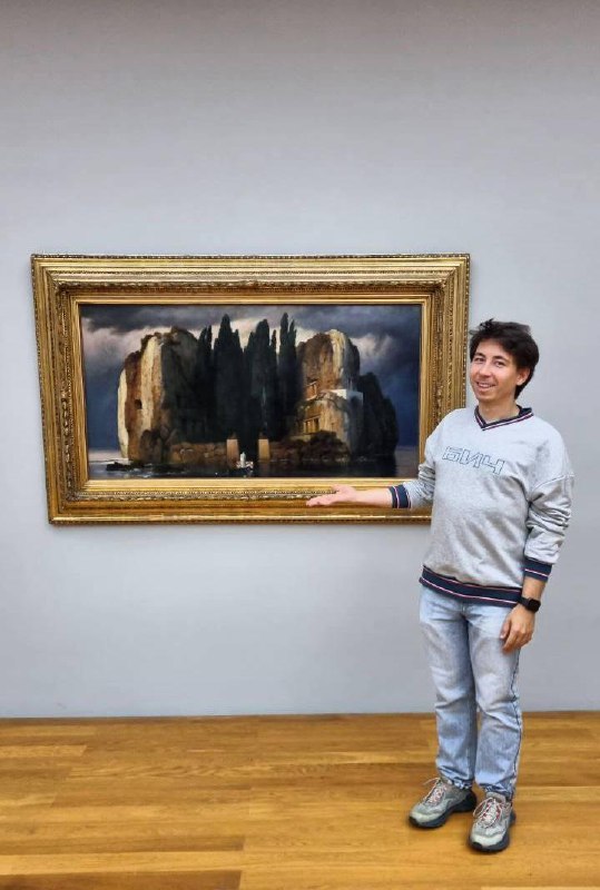 Человек позирует рядом с рамой и репродукцией «Острова мёртвых» в музейной зале, показывая масштаб картины и интерьер галереи.