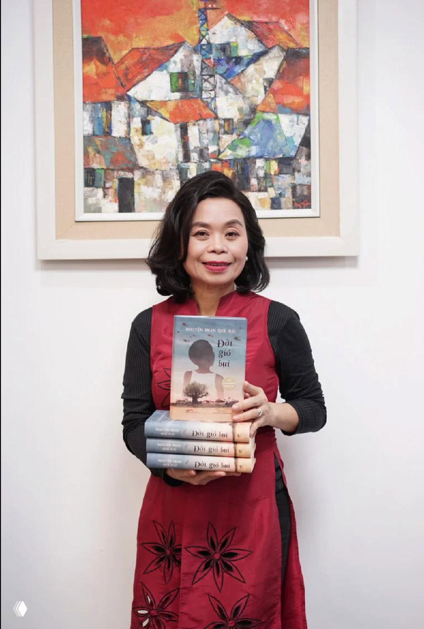 Портретная фотография авторки Нгуен Фан Кюэ Май: улыбающаяся женщина держит стопку своих книг на фоне живописи в интерьере.