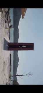 Инсталляция на берегу: вертикальная скульптура на фоне моря и гор, минималистичная композиция, сочетающая ландшафт и художественный объект.