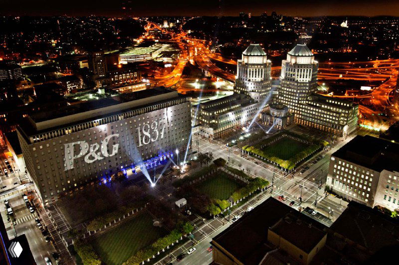 Ночное проецирование логотипа P&G на фасады зданий и освещённая городская сцена — пример массовой визуальной коммуникации и брендинга на уровне корпораций.