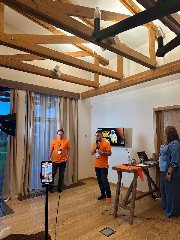 Интерьер кэмпа: два выступающих в оранжевых футболках ведут короткие доклады в уютном помещении с деревянными балками и экраном.