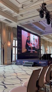 Конференц-зал с большим экраном, на экране показывают кадр превью видео о выступлении по ИИ; в кадре видны сцена, ряды кресел и оформление зала