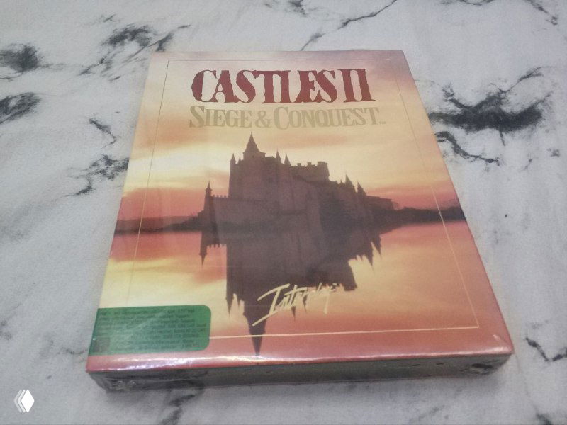 Запечатанная коробка Castles II (Siege & Conquest) на мраморной поверхности; видна фронтальная обложка с изображением замка и логотипом игры.