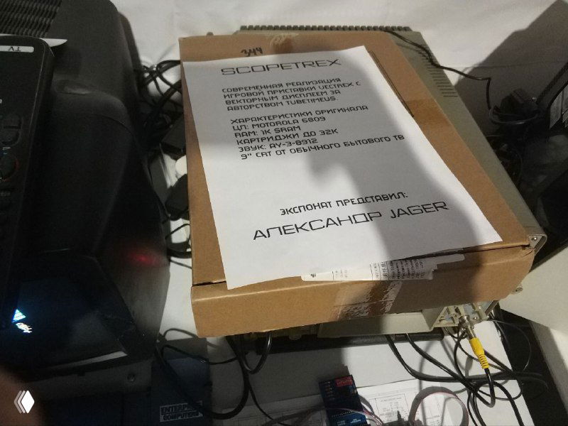 Коробка с сопроводительной бумажкой: лист с описанием имитации Vectrex или устройства лежит на коробке, рядом видны разъёмы и элементы демонстрации на столе.