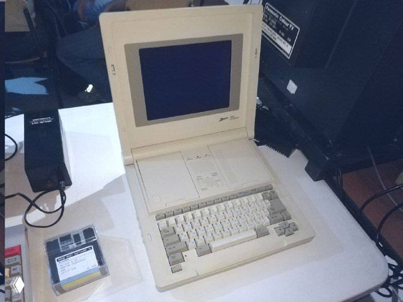 Винтажный ноутбук Zenith белого цвета на выставочном столе с закрытым экраном, рядом картридж и кабели; фото показывает конструкцию и корпус устройства.